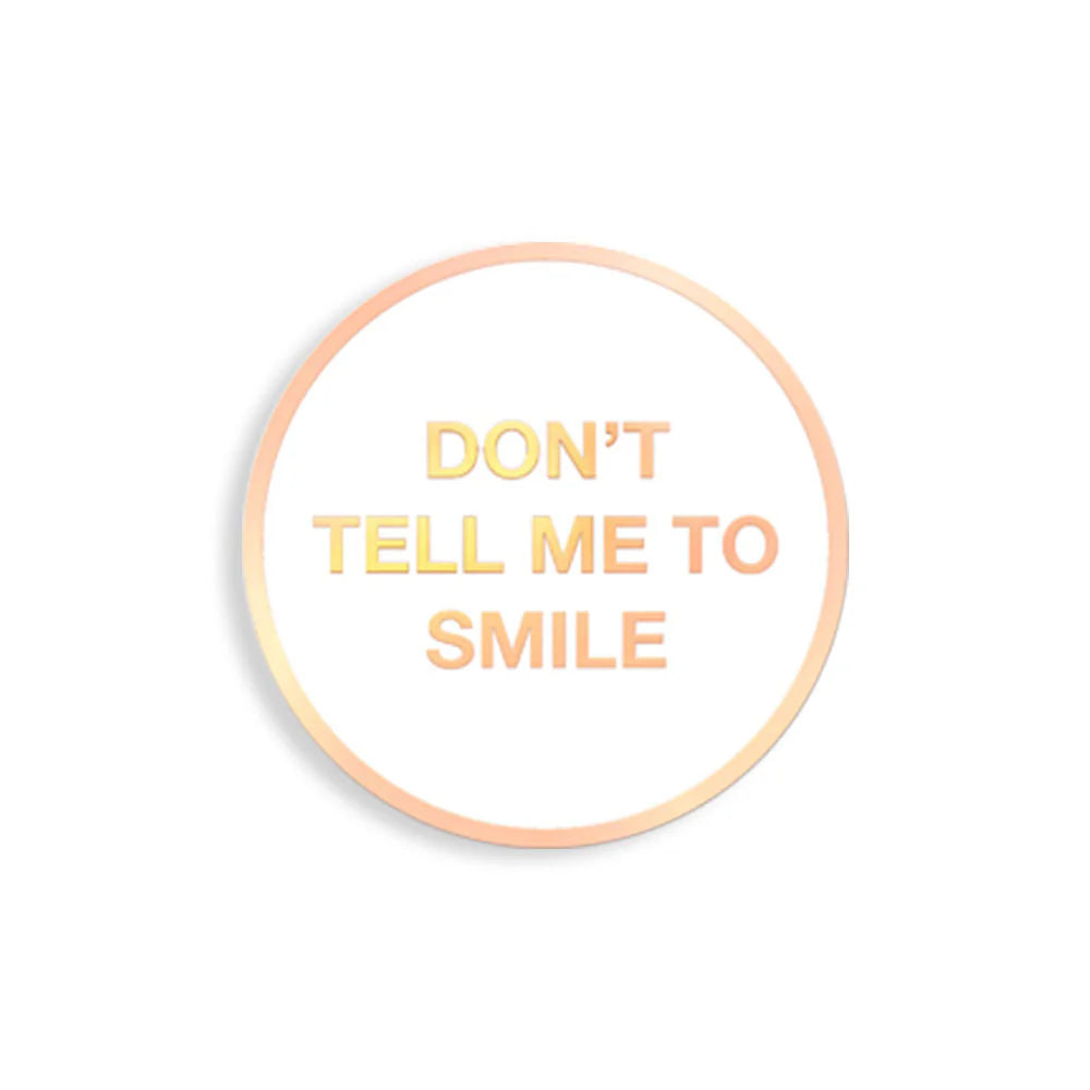 Round enamel pin with “Don’t Tell Me To Smile” in gold text