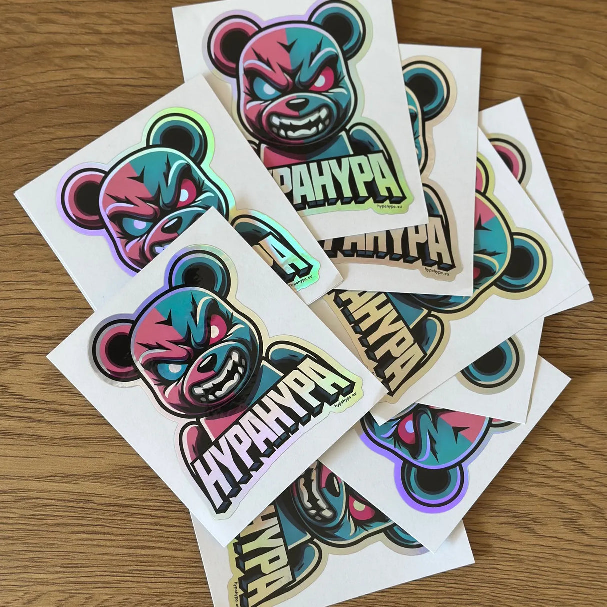 Ten HypaHypa holographic bear logo stickers spread out on a wooden table, showing colorful reflective shine.