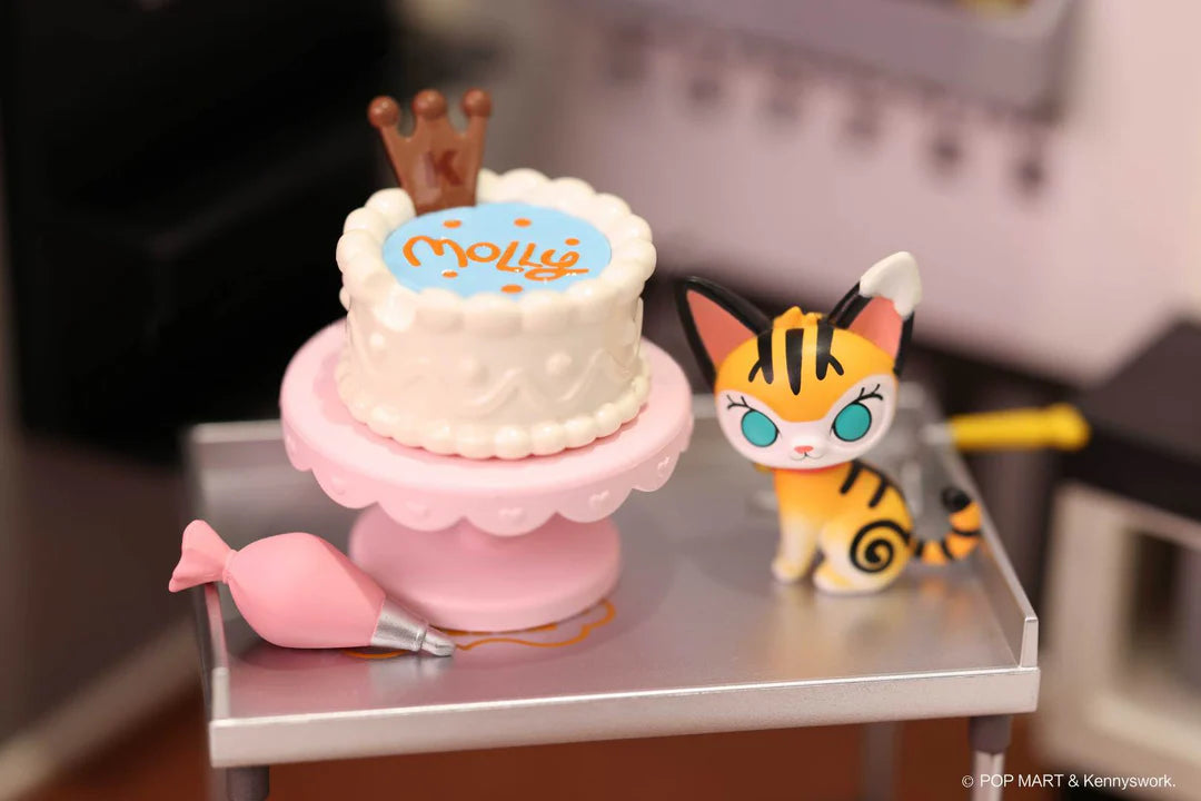 Cat figure standing beside white cake on pink tray with “Molly” written on top in icing