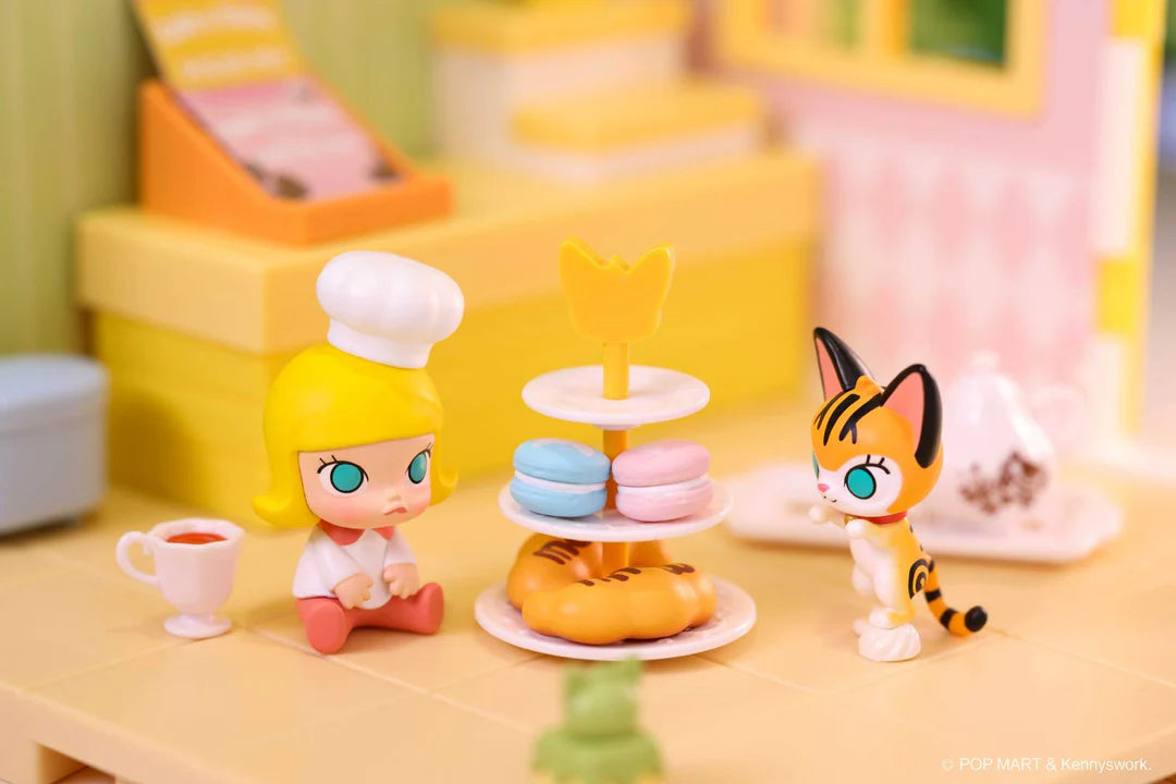 Molly chef figure with macarons and cat figure around a tiered dessert tray in pastel kitchen setting
