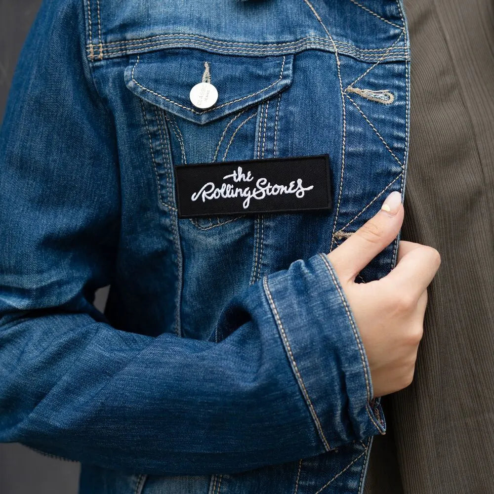 Close-up of a denim jacket with The Rolling Stones embroidered script logo patch on the pocket.