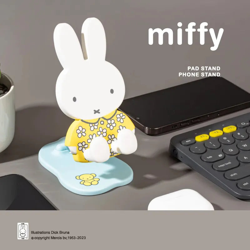 Miffy Folding Phone Stand Series figure with yellow floral outfit displayed on a desk beside a smartphone and keyboard
