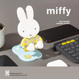 Miffy Folding Phone Stand Series figure with yellow floral outfit displayed on a desk beside a smartphone and keyboard