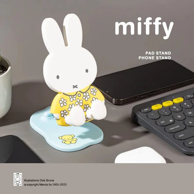 Miffy Folding Phone Stand Series figure with yellow floral outfit displayed on a desk beside a smartphone and keyboard